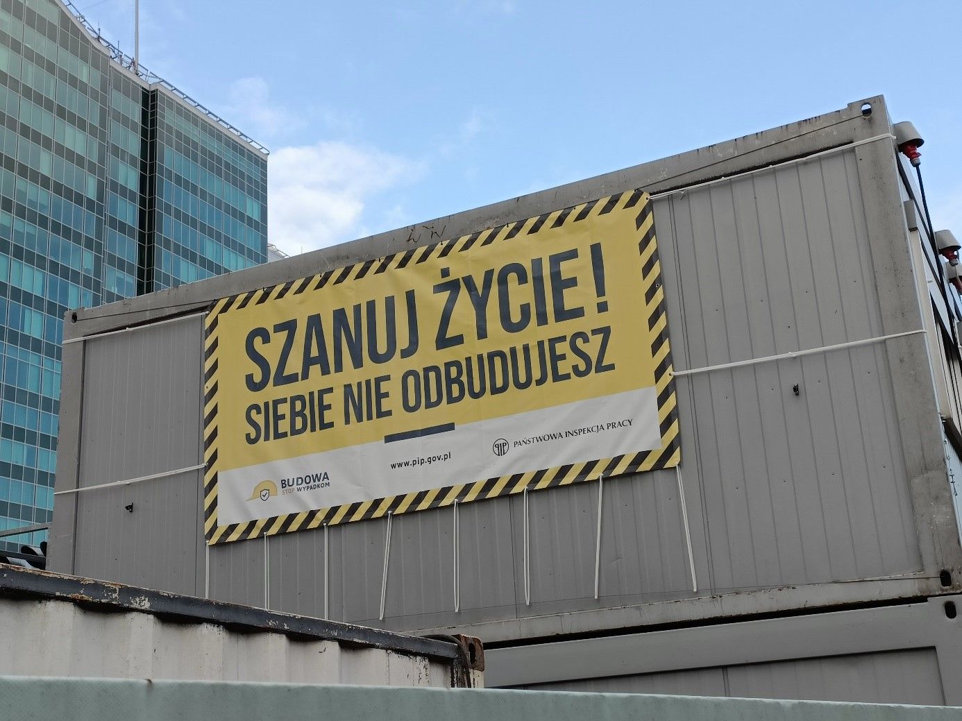 Na zdjęciu widać baner z napisem "Szanuj życie! Siebie nie odbudujesz". Baner zawieszony jest na ścianie kontenera mieszkalnego dla robotnoków budowlanych